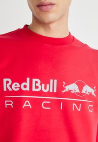Camiseta roja de algodón con un gran logo blanco de "Red Bull" y el texto "RACING" en una fuente moderna y audaz. Diseño de cuello redondo.
