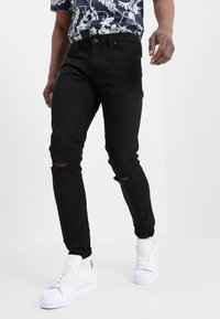 Svarta slitna skinny jeans med revor vid knäna, kombinerade med en blommönstrad skjorta och vita sneakers med snäckformad tådesign.