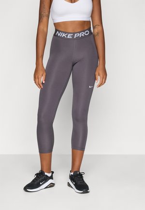 Gray Nike Pro leggings with a thick waistband featuring "NIKE PRO" in white. The leggings are fitted and extend to mid-calf. Black athletic shoes.