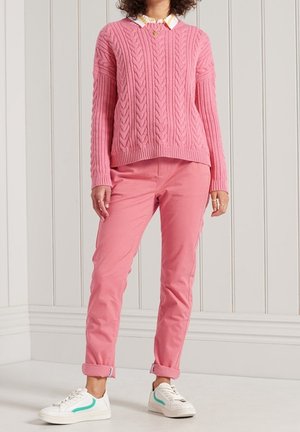 Personne portant un pull rose en maille torsadée, un pantalon rose retroussé, des baskets blanches avec des accents turquoise, debout contre un mur blanc à pans.