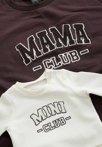 Tumši brūns treniņjak ar baltām teksturētām burtiem "MAMA CLUB" virs gaismas krēma treniņjaka ar brūnām burtiem "MINI CLUB".