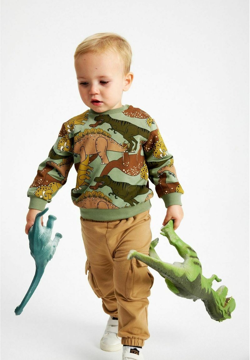Bambino che indossa una felpa verde con motivo di dinosauri e pantaloni cargo beige, tiene in mano dei giocattoli di dinosauri blu e verdi, mentre cammina su uno sfondo neutro.