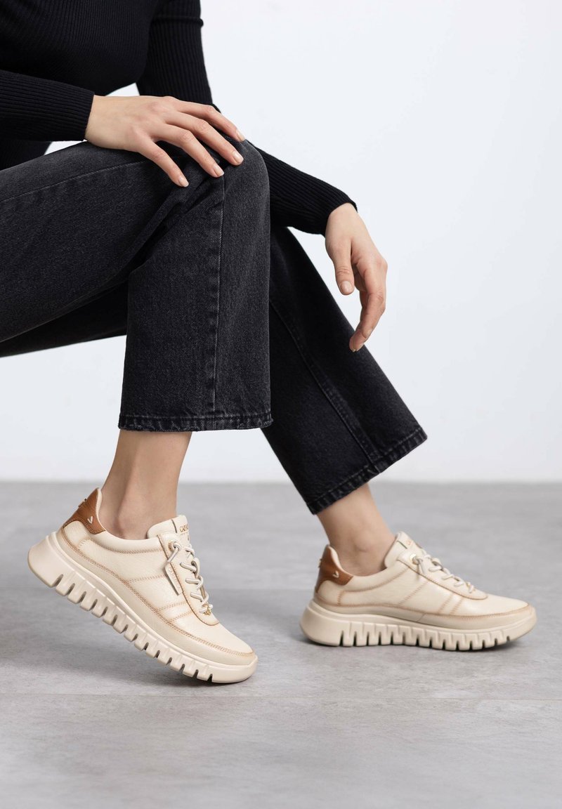 Zapatillas beige con superficie texturizada, acentos marrones en el talón, diseño con cordones y suela blanca gruesa con patrón estriado.