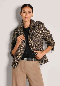 Vrouw in een jasje met luipaardprint, zwarte shirt, beige broek en ringoorbellen, poserend tegen een beige muur.