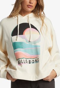 Hoodie color crema con una grande grafica che rappresenta un tramonto sopra le onde, con il testo "BILLA BONG" in basso.