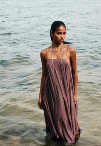 Robe mauve fluide sans manches avec une coupe décontractée, dotée de fines bretelles et d'un ourlet volé, portée au-dessus de l'eau avec des colliers en argent superposés.