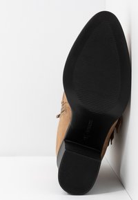 Bottine beige en daim avec une semelle noire en caoutchouc. Dotée d'un bout rond et d'une surface texturée, elle est rehaussée de petits détails décoratifs.