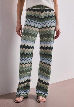 Ben iført højttaljede strikkede bukser med grønne, blå, beige og sorte zigzag-mønstre, kombineret med sølvfarvede sandaler og en hvid top.