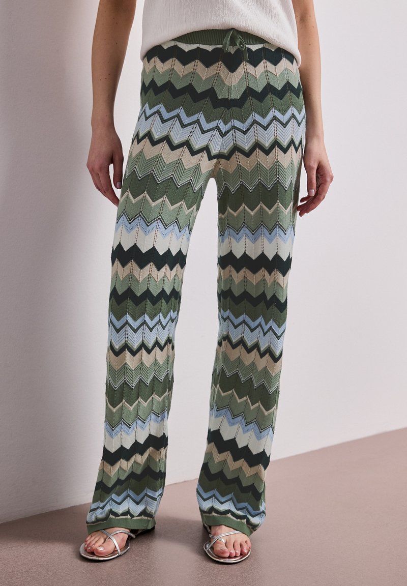 Jambes portant un pantalon en maille taille haute avec des motifs en zigzag verts, bleus, beiges et noirs, associé à des sandales argentées et un haut blanc.