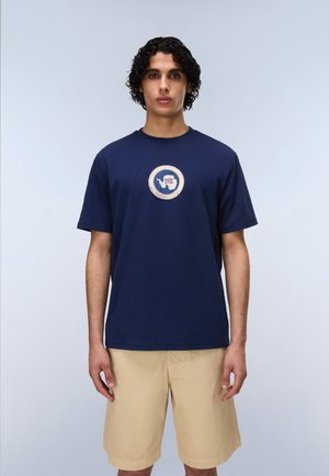 Młody mężczyzna o kręconych włosach, ubrany w granatowy t-shirt z okrągłym logo i beżowe spodenki do kolan, stojący na jednolitym tle.