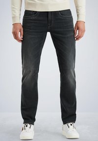 Muž v vyblednutých čiernych džínsach a bielych teniskách stojí proti jednému svetlošedému pozadiu.