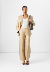 Costume en lin beige avec une veste sur mesure, un pantalon large assorti et un haut ajusté blanc. Portant un sac à main gris foncé. Escarpins noirs à lanières.