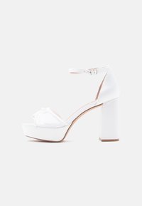 Λευκό σανδάλι με ψηλό τακούνι και πλατφόρμα, λουράκι στον αστράγαλο και διακοσμητικό φιόγκο στο μπροστινό μέρος. Ομαλό ύφασμα και τετράγωνο χοντρό τακούνι.