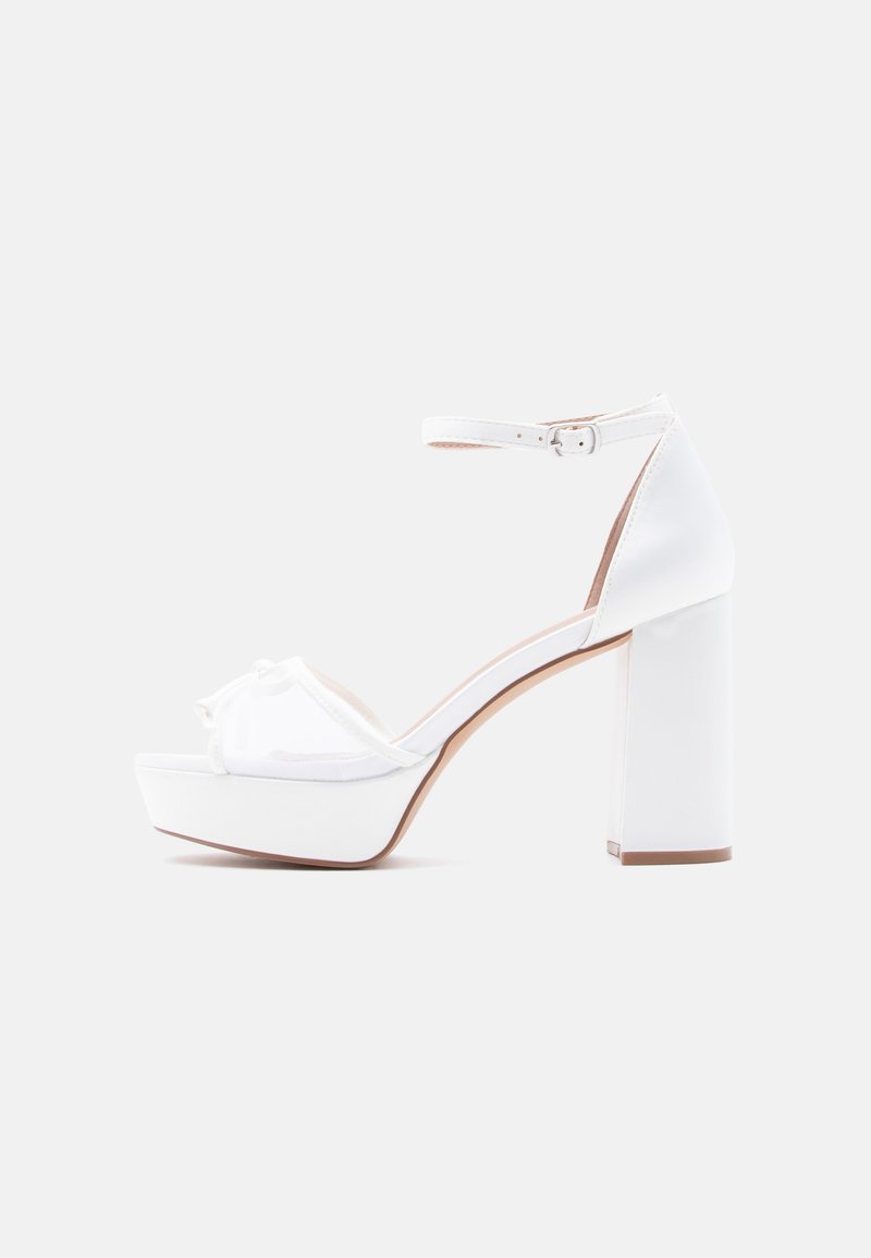 Λευκό σανδάλι με ψηλό τακούνι και πλατφόρμα, λουράκι στον αστράγαλο και διακοσμητικό φιόγκο στο μπροστινό μέρος. Ομαλό ύφασμα και τετράγωνο χοντρό τακούνι.