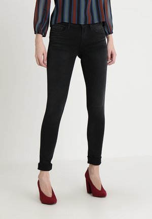 Femme portant un jean skinny noir retroussé à la cheville, des talons hauts rouges et un chemisier bleu rayé à manches longues, debout sur un sol blanc.