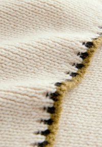 Strickstoff in Cremefarbe mit einem strukturierten Muster, das am Rand mit einem kräftigen Schwarz- und Goldakzent versehen ist. Weiche, dicke Garnstruktur.