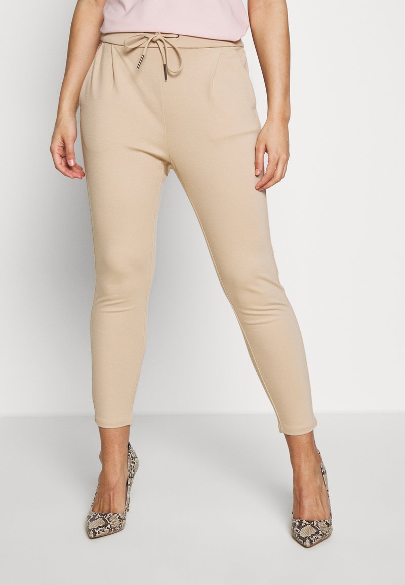 Kvinna i beige smala byxor med dragsko i midjan och högklackade skor med ormskinnsmönster, stående mot en vit bakgrund.