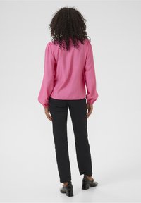 Blouse rose à manches longues avec épaules bouffantes, associée à un pantalon noir ajusté. La blouse a une texture lisse et une coupe décontractée.