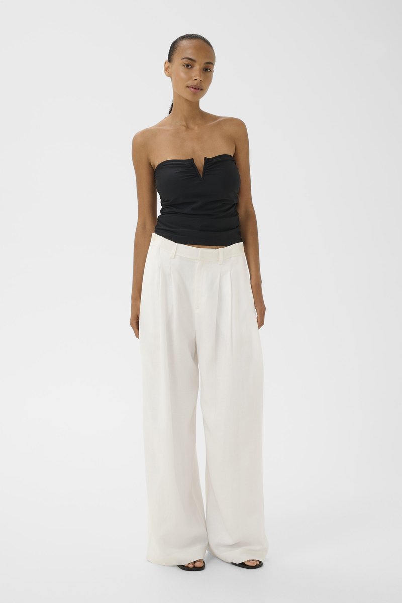 Frau steht und trägt ein trägerloses schwarzes Top und weiße, weit geschnittene Hosen mit offenen schwarzen Sandalen vor hell einfarbigem Hintergrund.