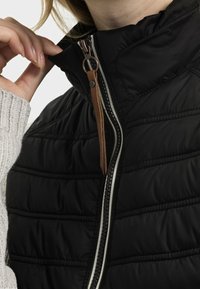 Svart puffig väst med hög krage, har en kontrasterande vit dragkedja och ett dragkedjehandtag i brunt läder, texturerade quiltade detaljer.