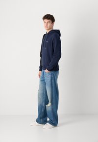 Felpa di cotone blu navy con tasca a marsupio e cappuccio regolabile, abbinata a jeans a gamba larga in denim chiaro strappato e sneakers bianche.