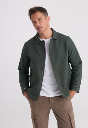 Man with hands in pockets wearing dark green zip jacket, white t-shirt, and brown pants against a plain light background.