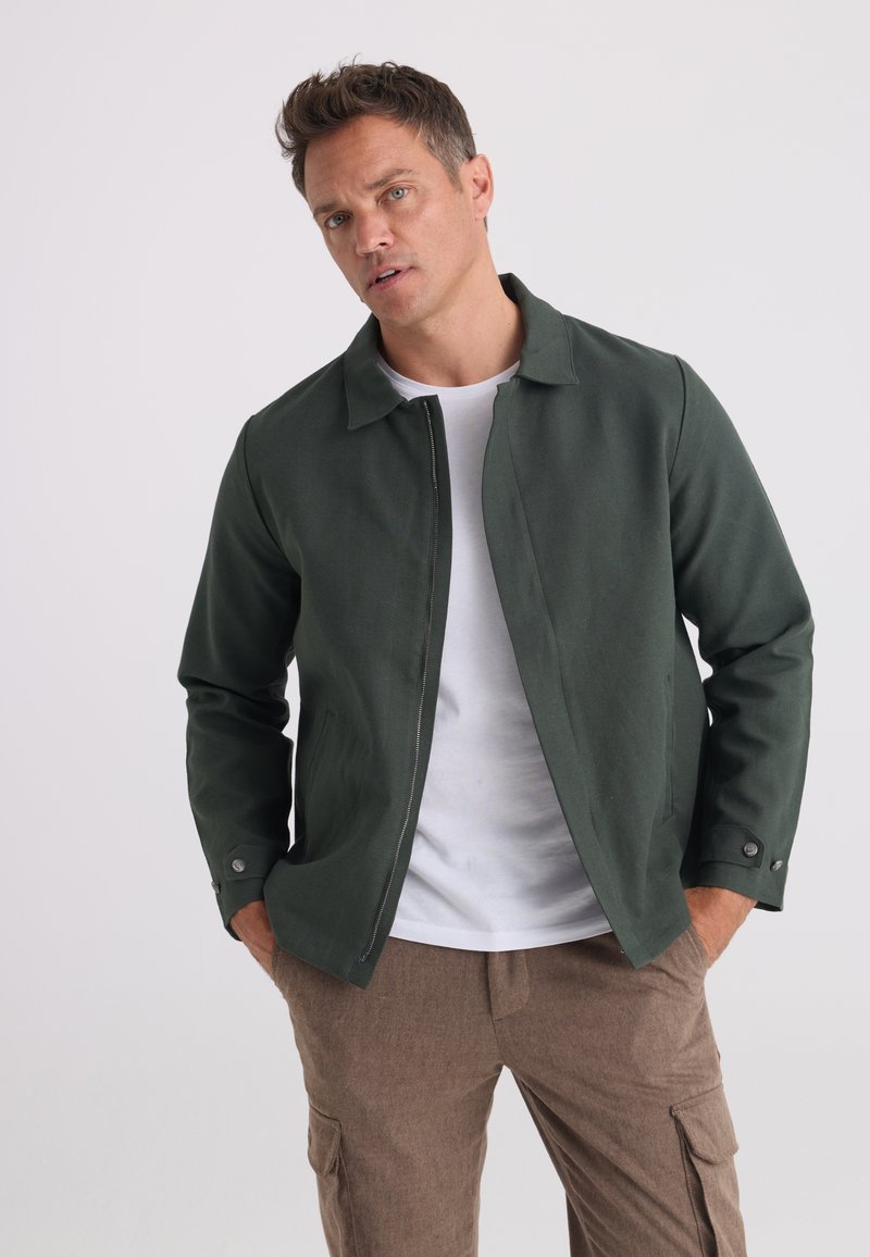 Homme les mains dans les poches portant une veste zippée vert foncé, un t-shirt blanc et un pantalon marron sur un fond clair uni.