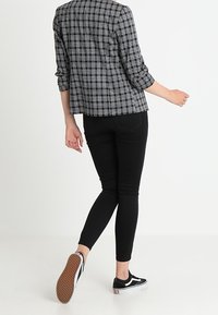 Osoba ubrana w czarno-białą kurtkę w kratę, czarne dopasowane spodnie i czarne sneakersy, idąca z uniesioną do tyłu jedną nogą.