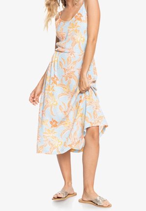 Vrouw draagt een mouwloze lichtblauwe jurk met oranje bloemenprint, houdt de rok vast en draagt zilveren platte sandalen.