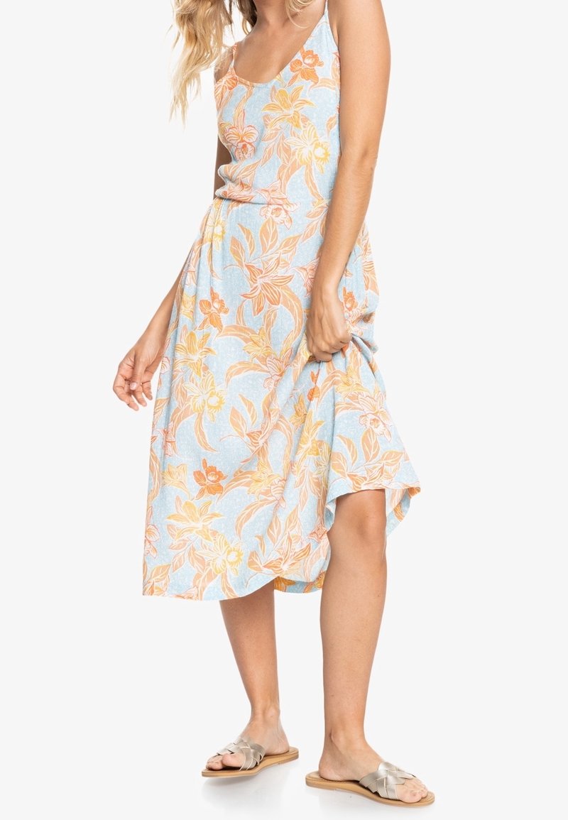 Femme portant une robe sans manches bleu clair avec un imprimé floral orange, tenant la jupe et portant des sandales plates argentées.