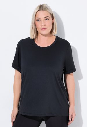 Blondynka w czarnej koszulce z krótkim rękawem i czarnych legginsach, stojąca przy białej ścianie, zwrócona przodem z neutralnym wyrazem twarzy.