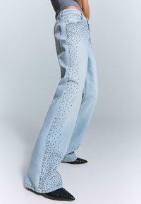 Light blue denim flared jeans with a studded pattern along the outer seams; features a zipper and button closure, paired with black pointed shoes.