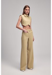 Beige cropped mouwloos topje met een puntige kraag en een knoopsluiting aan de voorkant, gecombineerd met hooggepolsterde, wijde broekspijpen in bijpassende stof.