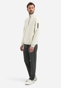 Lichtcrème half-zip pullover met een ritssluiting op de mouw, gecombineerd met donkergroene broeken en witte sneakers. Gladde textuur.