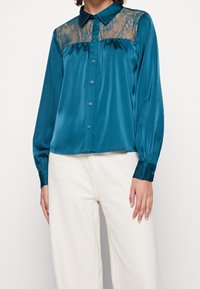 Tealfärgad satinblus med spetsdetaljer vid axlarna, knappstängning fram och långa ärmar med knappribbor. Slät textur och lös passform.