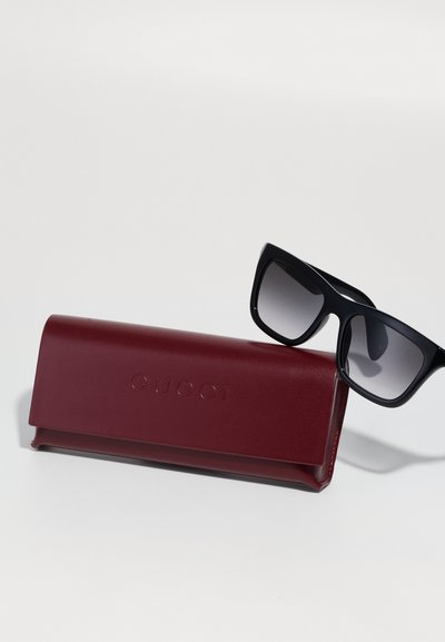 Čierne slnečné okuliare s gradientovými šošovkami spárované s bordovým tvrdým puzdrom s embosovaným logom "GUCCI". Puzdro má hladký povrch.