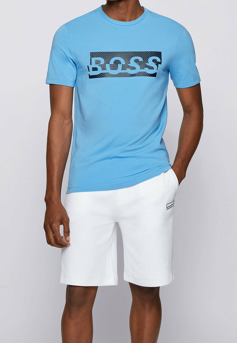 Homme portant un t-shirt bleu vif avec le texte "BOSS" et un short blanc jusqu'aux genoux, une main dans la poche.
