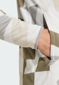 Flis jakna s geometrijskim uzorkom u bež, sivoj i maslinasto zelenoj boji. Uključuje bočnu džepu i sive manžete s mekom teksturom.