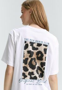 T-shirt bianco in cotone con una grafica leopardata in nero e beige sulla schiena, accompagnata dal testo "THE NEW DENIM CLUB" sopra di essa.