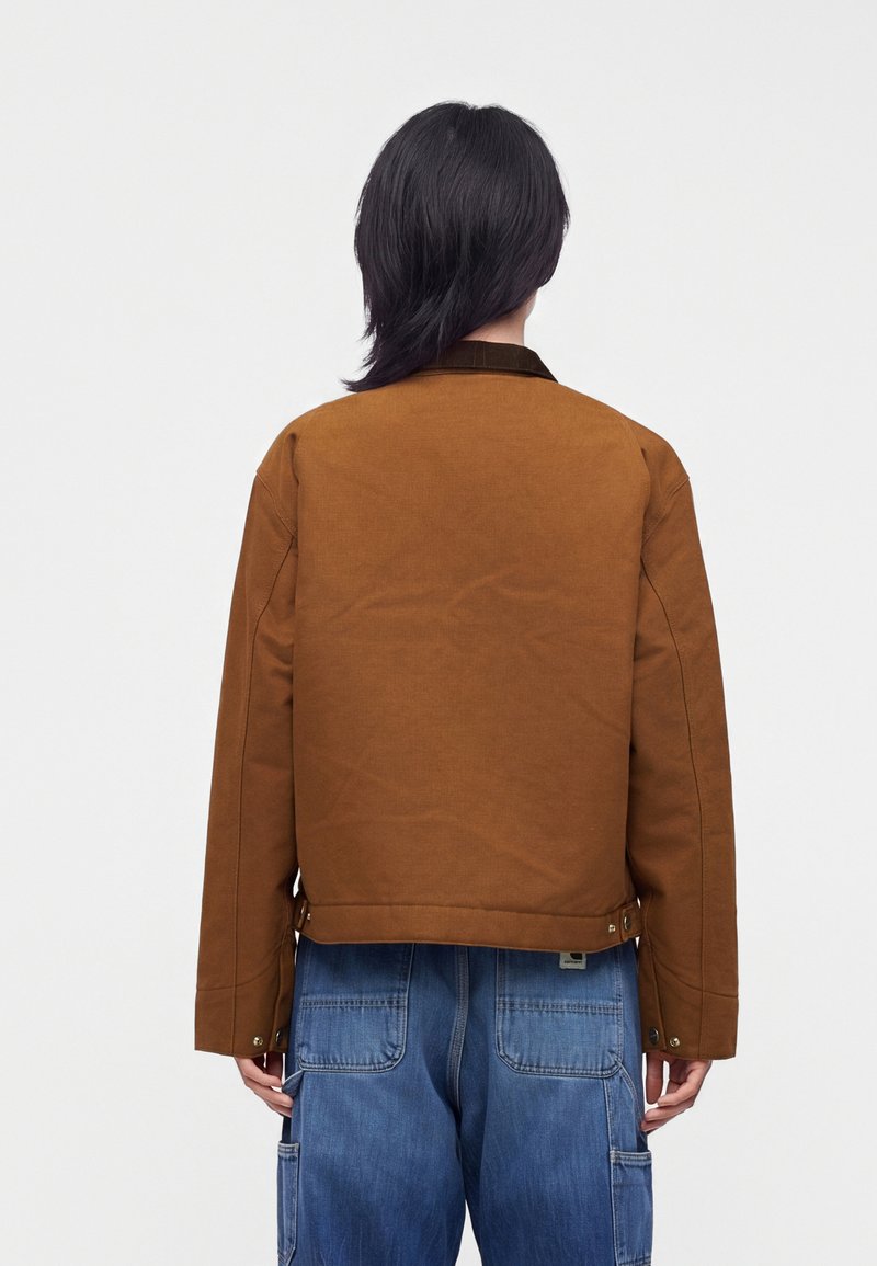 Person with black hair wearing a brown jacket and blue denim pants, standing with back facing the camera against a plain background.