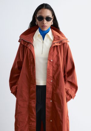 Kvinde iført oversized rust-rød frakke, cremefarvet lynlåstop over blåt rullekrave, sorte bukser, runde sorte solbriller og sølvfarvede creoløreringe.