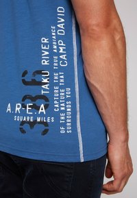 Camiseta azul con texto y gráficos impresos en blanco. Presenta un escote redondo, mangas cortas y costuras laterales en contraste.