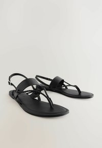 Sorte flade sandaler har flettede stropper, en justerbar ankelrem og en glat overflade. Designet er minimalistisk med en tåpost.