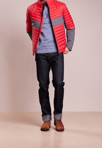 Red quilted jacket with grey panels, layered over a light blue shirt, paired with dark denim jeans and brown boots. Smooth texture, modern design.