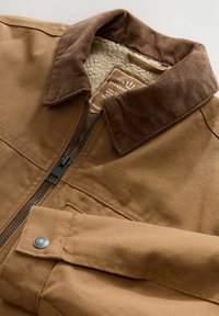 Chaqueta de lona marrón con cremallera y cuello de pana, forrada de felpa y con un detalle de botón a presión en la manga. Presenta costuras resistentes.