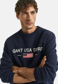 Navy blue cotton sweatshirt featuring "GANT USA SPRT" in white text and an embroidered American flag. Ribbed cuffs and crew neck design.