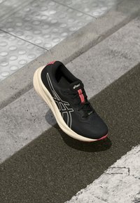 Chaussure de course noire avec une tige texturée, des accents blancs et roses, et une semelle rembourrée, posée sur une route avec un pavé texturé.