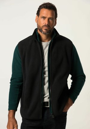 Chaleco de forro polar negro con interior de forro polar verde, cremallera frontal y dos bolsillos laterales. Lleva puesto sobre una camiseta gris, combinado con jeans oscuros.