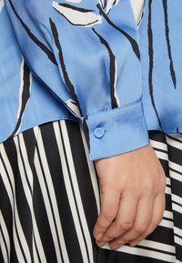 Mano appoggiata al fianco, indossa una camicia a maniche lunghe blu con motivo e una gonna a strisce verticali bianco e nere.