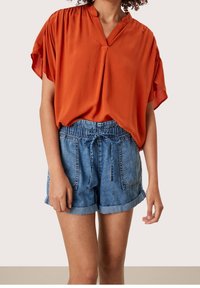 Orange relaxed-fit blouse with short, wide sleeves and a v-neck, paired with light blue denim shorts featuring an elastic waistband and pockets.